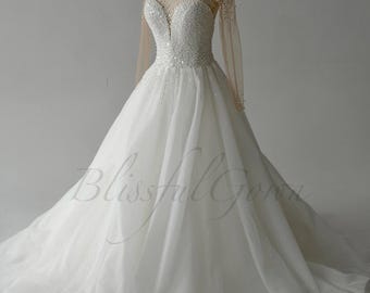 May include: A white wedding gown with a sweetheart neckline and long, sheer sleeves. The bodice is embellished with sparkling beads, and the skirt flows into a full, layered train. The dress is displayed on a mannequin.