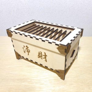 May include: A light-colored wooden box with a slatted top and decorative corner pieces. The box has Japanese characters on the front. The box is made of light-colored wood with darker brown accents.