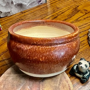 May include: A handmade, round ceramic bowl with a wide rim. The bowl is glazed in shades of brown and tan, with a speckled texture. The interior is a lighter tan color. The bowl sits on a white base. A small panda figurine is next to the bowl.
