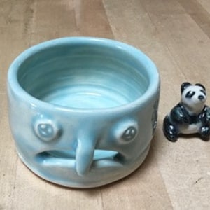 May include: A light blue ceramic bowl with a face design, featuring a nose, eyes, and a mouth. Beside it is a small panda figurine in black and white. The bowl is on a light wood surface.