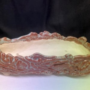 May include: A rectangular ceramic planter with a textured, reddish-brown exterior and a light interior. The rim has a wavy design with a hint of blue-green. The planter is suitable for small plants or succulents.