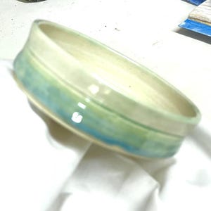 May include: A shallow, round ceramic bowl with a smooth, cream-colored interior. The exterior features a blended glaze of soft green and blue tones, creating a horizontal gradient effect around the curved sides of the vessel.