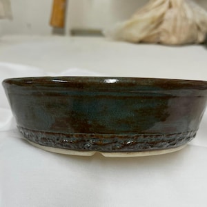May include: A shallow, round ceramic bowl with a glossy, dark brown and deep teal glaze. The exterior features a textured, rugged band around the lower portion, while the base is a contrasting light, unglazed off-white ceramic material.