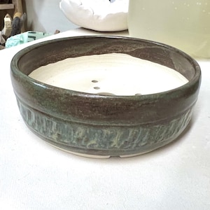 May include: A handmade ceramic bowl with a textured, earthy design. The bowl features a wide, shallow shape with a white interior and a rim in shades of green and brown. The exterior has a textured band around the middle.