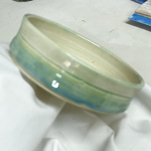 May include: A shallow, round ceramic bowl with a smooth, cream-colored interior. The exterior features a blended glaze transition from a soft, pale green at the top rim to a vibrant, deep turquoise blue at the base, creating a gradient effect.