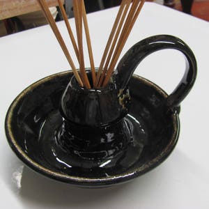 May include: A black ceramic incense holder with a handle and a small bowl. The holder has a glossy finish with a gold rim and holds several wooden incense sticks. The design is simple and elegant.