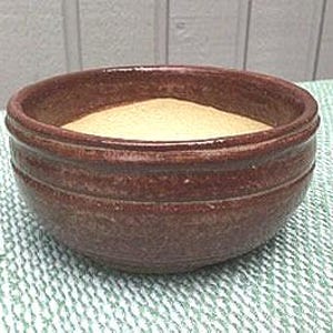 May include: A small, round, brown ceramic bowl filled with a light tan substance. The bowl has a textured, ridged exterior and sits on a green and white patterned surface. The bowl is centered in the image.