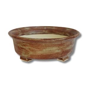 May include: A round, brown ceramic bonsai pot with a light interior. The pot has a textured surface and is supported by three small, square feet. The rim is slightly flared, and the overall design is simple and functional.