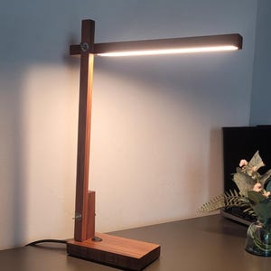 Op de afbeelding: Een houten bureaulamp met een rechthoekige basis en een verticale steun. De lamp heeft een horizontale lichtbalk aan de bovenkant. Het hout heeft een warme bruine kleur en de lamp is ingeschakeld en geeft helder licht af.