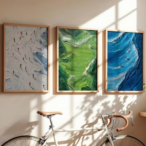 May include: Three framed art prints with wooden frames hang on a beige wall. The prints depict a snowy mountain scene with skiers, a green landscape, and a blue ocean wave with surfers. A bicycle is in the foreground.