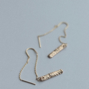 May include: A pair of gold-colored threader earrings. Each earring features a delicate chain attached to a textured, rectangular bar with an engraved floral design. The earrings are displayed against a light blue background.