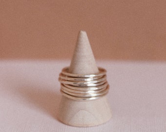May include: A stack of silver rings on a white cone-shaped ring holder. The rings are all different sizes and have a hammered texture.