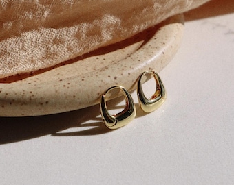 May include: A pair of gold-colored hoop earrings with a unique, rounded rectangular shape. The earrings are displayed on a beige, speckled ceramic plate, with a soft, beige fabric in the background. The earrings have a polished, reflective surface.