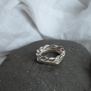 K&ouml;nnte beinhalten: Ein silberfarbener Ring mit einer quadratischen Basis, die mit einem gedrehten, seilartigen Metalldesign verziert ist. Der Ring hat eine polierte, reflektierende Oberfl&auml;che und besteht aus einem silberfarbenen Metall mit geometrischer Form.