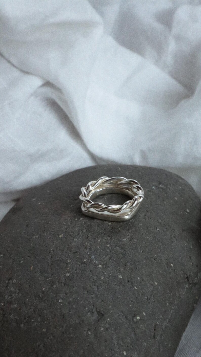 K&ouml;nnte beinhalten: Ein silberfarbener Ring mit einer quadratischen Basis, die mit einem gedrehten, seilartigen Metalldesign verziert ist. Der Ring hat eine polierte, reflektierende Oberfl&auml;che und besteht aus einem silberfarbenen Metall mit geometrischer Form.