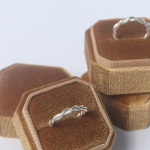 K&ouml;nnte beinhalten: Zwei silberne Ringe mit gedrehtem Seildesign, die jeweils auf einer separaten achteckigen Ringbox aus braunem Samt liegen. Die Ringe haben eine polierte metallische Oberfl&auml;che und ein durchgehendes Flechtmuster um das gesamte Band.