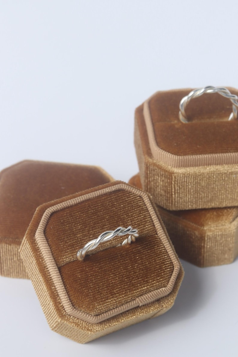 K&ouml;nnte beinhalten: Zwei silberne Ringe mit gedrehtem Seildesign, die jeweils auf einer separaten achteckigen Ringbox aus braunem Samt liegen. Die Ringe haben eine polierte metallische Oberfl&auml;che und ein durchgehendes Flechtmuster um das gesamte Band.