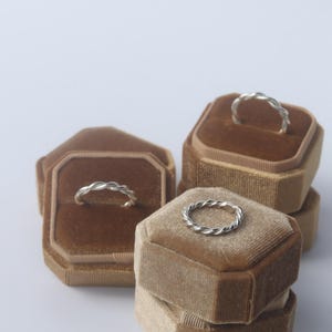 K&ouml;nnte beinhalten: Drei silberne Ringe mit gedrehter Seilstruktur, die jeweils auf einer achteckigen Ringbox aus braunem Samt pr&auml;sentiert werden. Die Ringe verf&uuml;gen &uuml;ber ein durchgehendes geflochtenes Metalldesign mit polierter Oberfl&auml;che und runder Form.