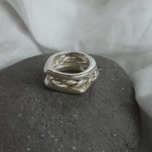 K&ouml;nnte beinhalten: Ein silberner Ring mit einem gestapelten Design aus zwei verschiedenen B&auml;ndern. Das obere Band besteht aus einem glatten, abgerundeten Silberdraht, w&auml;hrend das untere Band aus einem gedrehten, geflochtenen Silberdraht gefertigt ist. Polierte Oberfl&auml;che.