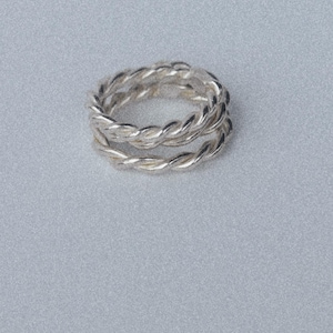 K&ouml;nnte beinhalten: Ein silberner Ring mit gedrehtem Seildesign. Das Band besteht aus zwei getrennten, ineinander verschlungenen Str&auml;ngen aus poliertem Silbermetall, die eine durchgehende, texturierte und kreisf&ouml;rmige Form mit einer reflektierenden Oberfl&auml;che bilden.