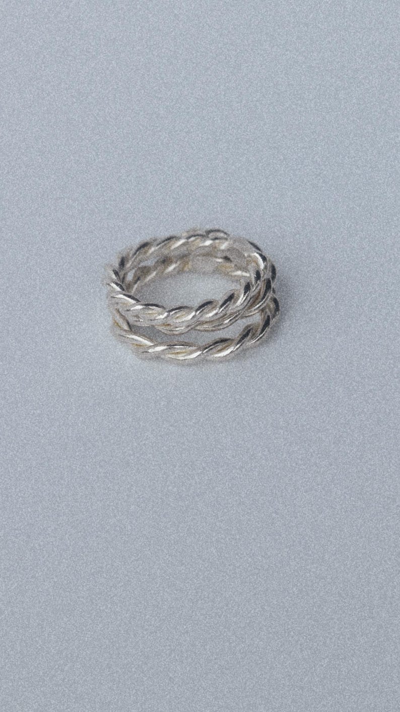 K&ouml;nnte beinhalten: Ein silberner Ring mit gedrehtem Seildesign. Das Band besteht aus zwei getrennten, ineinander verschlungenen Str&auml;ngen aus poliertem Silbermetall, die eine durchgehende, texturierte und kreisf&ouml;rmige Form mit einer reflektierenden Oberfl&auml;che bilden.