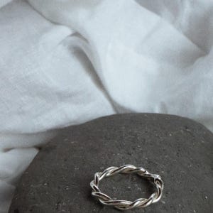 K&ouml;nnte beinhalten: Ein silberfarbener Ring mit einem gedrehten, seilartigen Design. Das Band besteht aus mehreren d&uuml;nnen Str&auml;ngen, die miteinander verflochten sind, um eine durchgehende, texturierte Kreisform mit einer polierten, reflektierenden Oberfl&auml;che zu bilden.