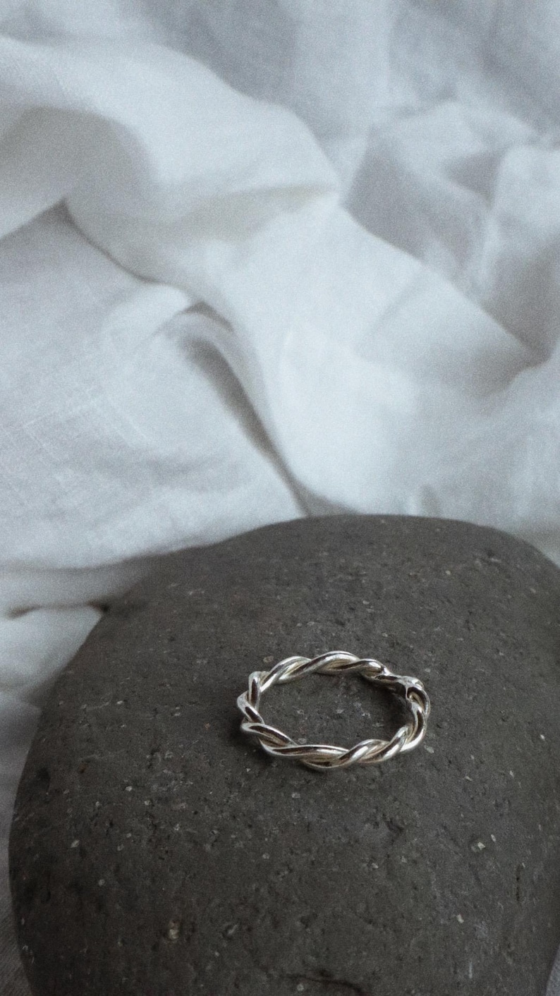 K&ouml;nnte beinhalten: Ein silberfarbener Ring mit einem gedrehten, seilartigen Design. Das Band besteht aus mehreren d&uuml;nnen Str&auml;ngen, die miteinander verflochten sind, um eine durchgehende, texturierte Kreisform mit einer polierten, reflektierenden Oberfl&auml;che zu bilden.
