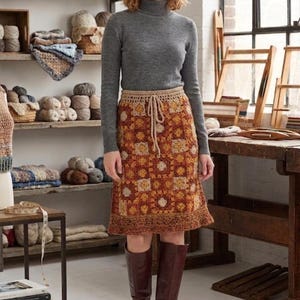 Peut inclure: Une personne porte un pull à col roulé gris et une jupe en crochet avec une taille à cordon. La jupe présente un motif patchwork dans les tons orange, marron et crème. Elle porte également des bottes hautes marron. Des étagères en arrière-plan présentent de la laine et des objets artisanaux tissés.