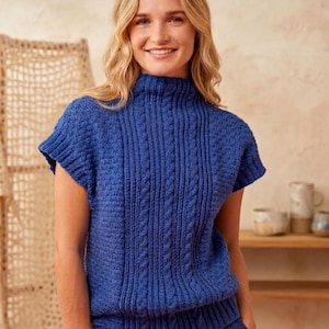 Peut inclure: Un haut bleu tricoté avec un col montant et des manches courtes. Le haut présente un motif de tricot torsadé sur le devant et un motif texturé sur les épaules. Le modèle porte un pantalon bleu. L'arrière-plan est de couleur neutre.