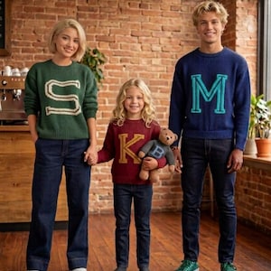 Peut inclure: Trois personnes portant des pulls assortis avec de grandes lettres initiales. Les pulls sont verts, bordeaux et bleus. Ils se tiennent devant un mur de briques et portent des jeans bleus. Une personne tient un ours en peluche.