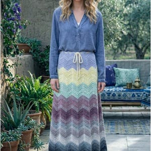 Peut inclure: Une jupe longue à motif chevron coloré avec une taille à cordon. Les couleurs de la jupe passent du bleu clair au jaune, vert, violet et gris. La personne porte également une chemise bleue à manches longues.