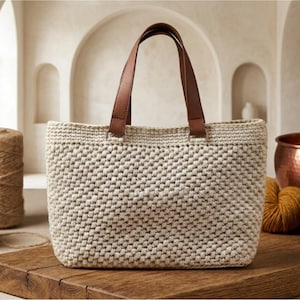 May include: A rectangular, off-white woven tote bag with brown leather handles. The bag is sitting on a wooden surface, with spools of twine and yarn in the background. A copper pot and yarn balls are also visible.