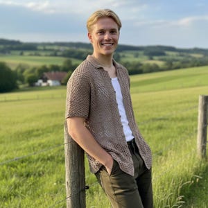 Peut inclure: Une chemise marron clair à tissage ouvert, à manches courtes et col. La chemise est boutonnée et portée sur un sous-vêtement blanc. La personne porte un pantalon vert foncé et s'appuie sur un poteau de clôture en bois dans un champ.