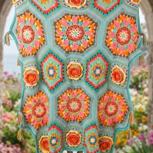 Peut inclure: Une couverture au crochet turquoise avec un motif floral dans les tons de rose, d'orange et de jaune. La couverture présente un motif géométrique avec des formes hexagonales et des glands décoratifs sur les bords. La couverture est tenue, mettant en valeur ses détails complexes.