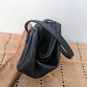 May include: A black leather crossbody bag with a top handle and a long adjustable strap. The bag has a textured surface and a zippered closure. The bag is held by a person.