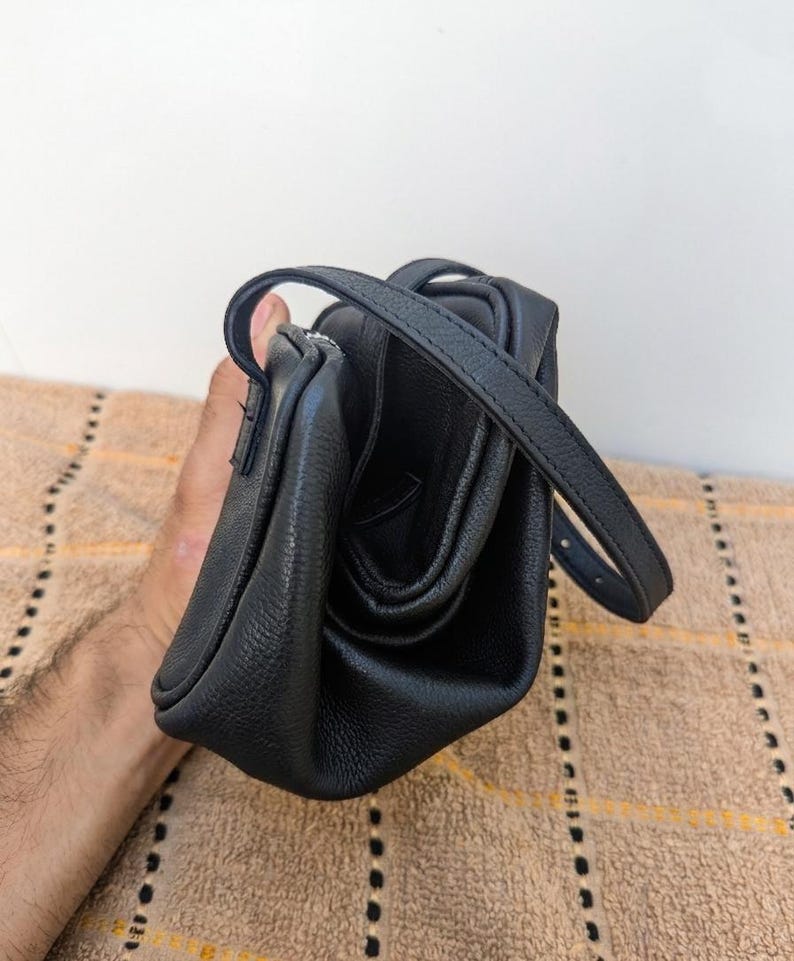 May include: A black leather crossbody bag with a top handle and a long adjustable strap. The bag has a textured surface and a zippered closure. The bag is held by a person.