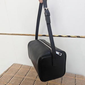 May include: Black rectangular handbag with a silver zipper and a matching adjustable strap. The bag is made of a smooth material and has a minimalist design. The strap is attached with a silver buckle.