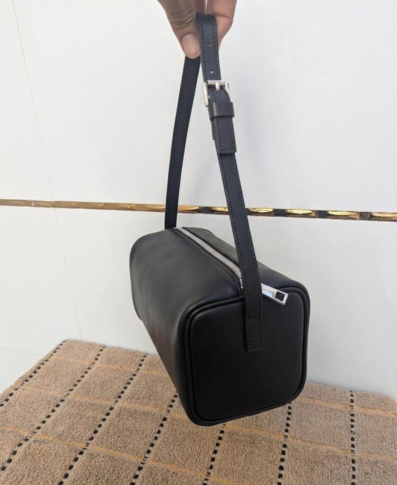 May include: Black rectangular handbag with a silver zipper and a matching adjustable strap. The bag is made of a smooth material and has a minimalist design. The strap is attached with a silver buckle.