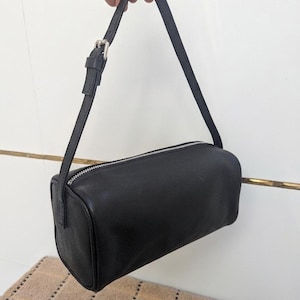 May include: A black, rectangular handbag with a silver zipper and a thin, adjustable strap. The bag is held up by a hand. The bag is approximately 10 inches long.