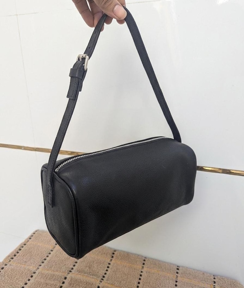 May include: A black, rectangular handbag with a silver zipper and a thin, adjustable strap. The bag is held up by a hand. The bag is approximately 10 inches long.