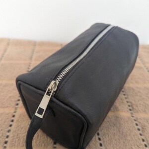May include: A black rectangular cosmetic bag with a silver zipper and a small handle. The bag is made of a smooth material and has a rectangular shape. The zipper is silver and runs along the top of the bag.