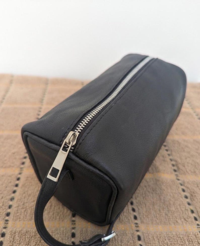 May include: A black rectangular cosmetic bag with a silver zipper and a small handle. The bag is made of a smooth material and has a rectangular shape. The zipper is silver and runs along the top of the bag.