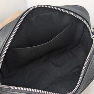 May include: Open view of a black leather crossbody bag with a silver zipper. The interior reveals a black fabric lining with a pocket. The bag is designed with a rectangular shape.