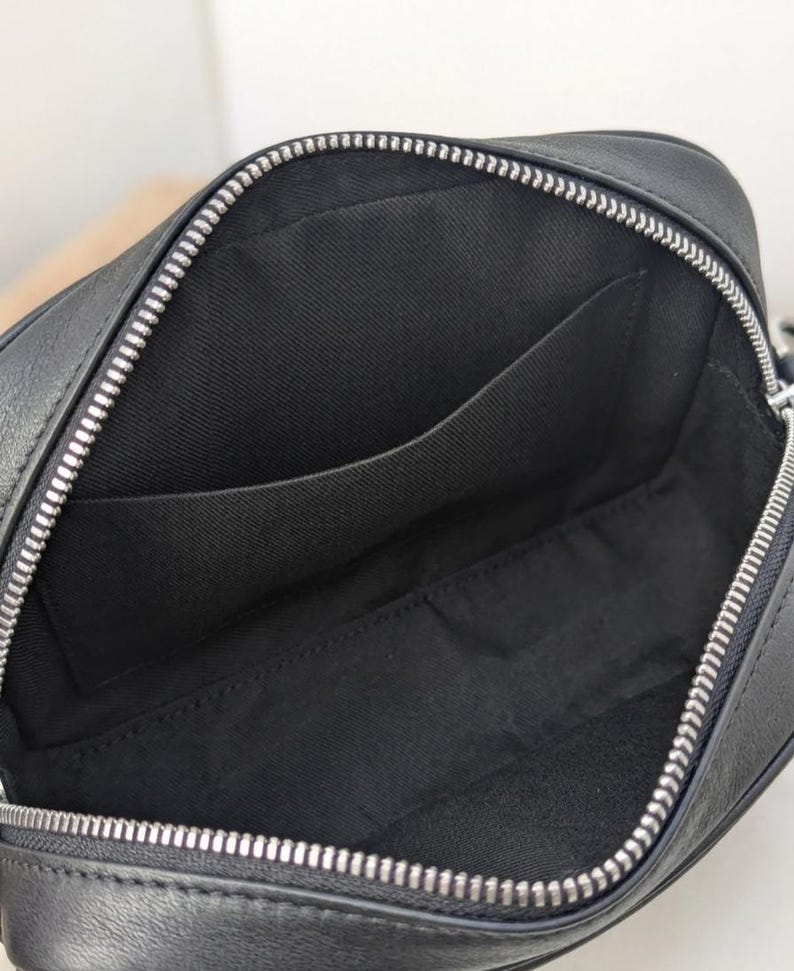 May include: Open view of a black leather crossbody bag with a silver zipper. The interior reveals a black fabric lining with a pocket. The bag is designed with a rectangular shape.