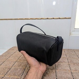 May include: A black, cylindrical handbag with a top handle and a zipper closure. The bag is made of a smooth material and is held by a person's hand. The bag is approximately 10 inches long.