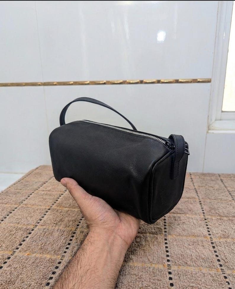 May include: A black, cylindrical handbag with a top handle and a zipper closure. The bag is made of a smooth material and is held by a person's hand. The bag is approximately 10 inches long.