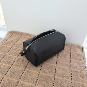 May include: A black rectangular toiletry bag with a silver zipper and a small handle. The bag is open, revealing the interior. The bag is made of a textured material and sits on a brown towel.