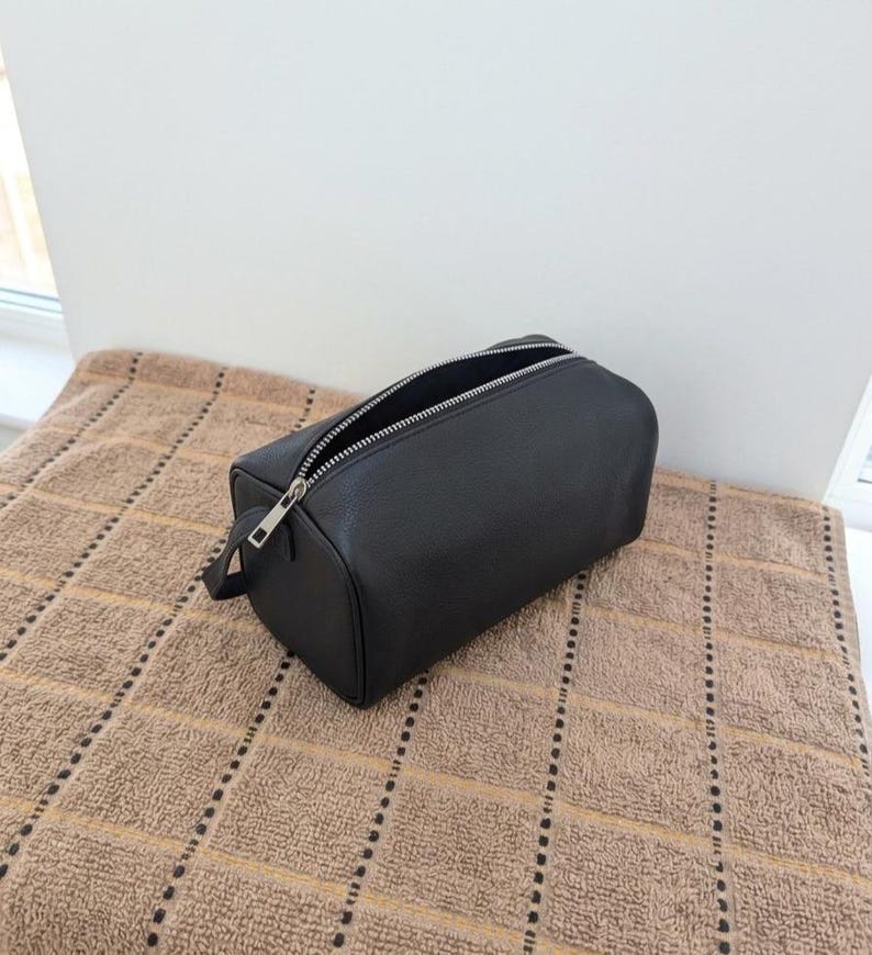 May include: A black rectangular toiletry bag with a silver zipper and a small handle. The bag is open, revealing the interior. The bag is made of a textured material and sits on a brown towel.