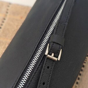 May include: Black leather bag with a silver zipper and buckle closure. The bag has a textured surface and a strap with a buckle. The zipper runs diagonally across the bag.