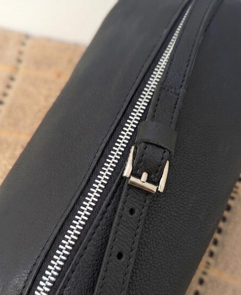 May include: Black leather bag with a silver zipper and buckle closure. The bag has a textured surface and a strap with a buckle. The zipper runs diagonally across the bag.